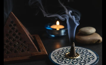 Incense cone burning on patterned holder with smoke, wooden incense box, candle, and stones