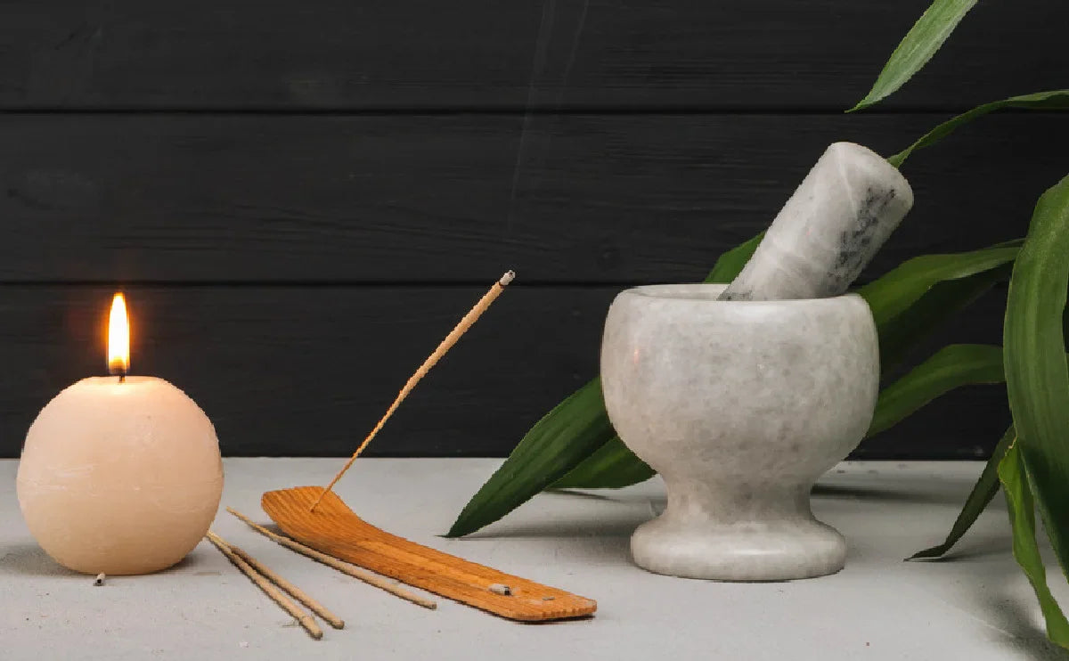Lit candle, burning incense stick on holder, and mortar and pestle on gray surface with green leaves