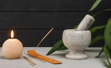 Lit candle, burning incense stick on holder, and mortar and pestle on gray surface with green leaves
